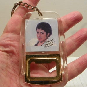 May include: A clear plastic keyring featuring a portrait of Michael Jackson with the text "Michael Jackson Thriller." The keyring has a gold-coloured bottle opener at the bottom and a metal ring for keys.