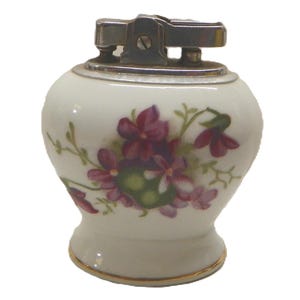 May include: A table lighter with a rounded white ceramic body featuring a painted floral pattern of purple flowers and green leaves. The top mechanism is made of polished silver-colored metal, and the base is accented with a thin gold-colored rim.
