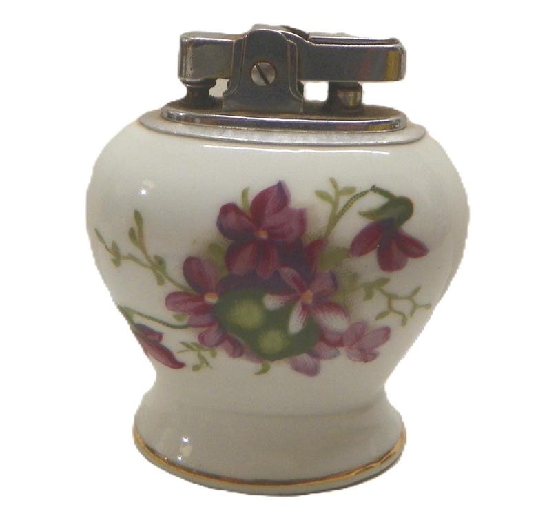 May include: A table lighter with a rounded white ceramic body featuring a painted floral pattern of purple flowers and green leaves. The top mechanism is made of polished silver-colored metal, and the base is accented with a thin gold-colored rim.