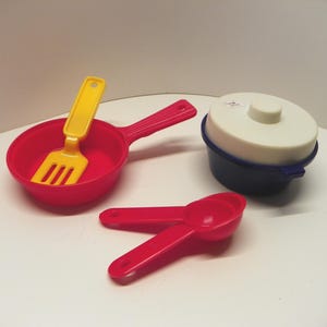 以下が含まれることがあります： カラフルなおもちゃのキッチン用品セット。赤いフライパンと黄色のヘラ、蓋付きの青と白の鍋、赤い計量スプーン2本が含まれています。おもちゃはプラスチック製です。