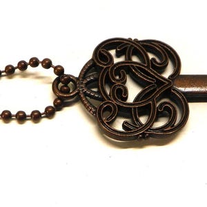 Op de afbeelding: Een bronskleurige metalen flesopener in de vorm van een sierlijke skelet-sleutel. De kop heeft een decoratief filigraanpatroon en de schacht is voorzien van een kogelketting. Het uiteinde is ontworpen met een functionele inkeping voor het openen van flessen.