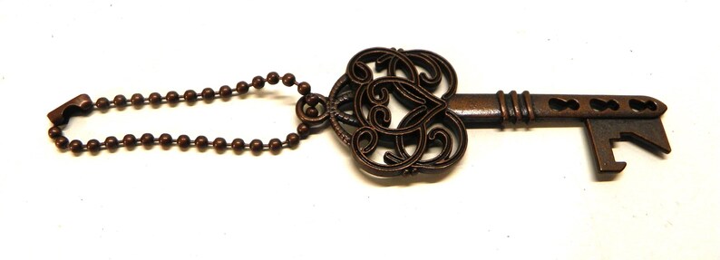 Op de afbeelding: Een bronskleurige metalen flesopener in de vorm van een sierlijke skelet-sleutel. De kop heeft een decoratief filigraanpatroon en de schacht is voorzien van een kogelketting. Het uiteinde is ontworpen met een functionele inkeping voor het openen van flessen.