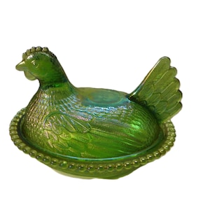 May include: A vibrant green iridescent glass hen on a nest dish. The lid is shaped like a sitting chicken with detailed feather textures, while the base features a decorative beaded rim. The glass has a reflective, metallic sheen throughout.