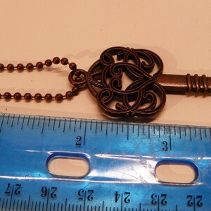 Op de afbeelding: Een bronskleurige metalen flesopener in de vorm van een sleutel met een sierlijke, filigraanachtige kop en een kogelketting. Een blauwe plastic liniaal ligt eronder en toont aan dat het object een totale lengte van ongeveer 11,5 cm heeft.