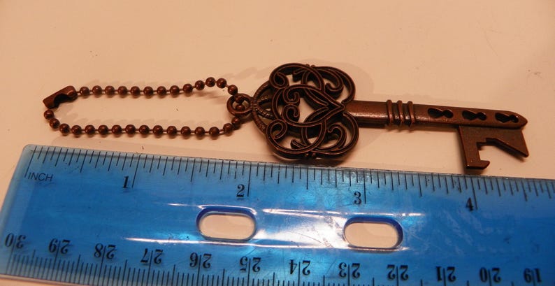Op de afbeelding: Een bronskleurige metalen flesopener in de vorm van een sleutel met een sierlijke, filigraanachtige kop en een kogelketting. Een blauwe plastic liniaal ligt eronder en toont aan dat het object een totale lengte van ongeveer 11,5 cm heeft.