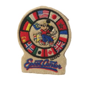 May include: An embroidered patch featuring Mickey Mouse in a red shirt and blue trousers, surrounded by a circular border of various national flags. Below the circle, the text Epcot Center is stitched in blue with a red outline on a beige background.