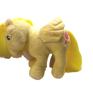 May include: A vintage plush toy pony in a pale yellow colour with yellow hair and wings. The pony has a pink heart-shaped mark on its flank. The toy is made of soft, plush material and has a friendly expression.