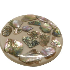 May include: A circular, beige decorative item featuring iridescent abalone shell fragments. The shells are of various sizes and shapes, showcasing green, pink, and silver hues. The object has a smooth, polished surface.