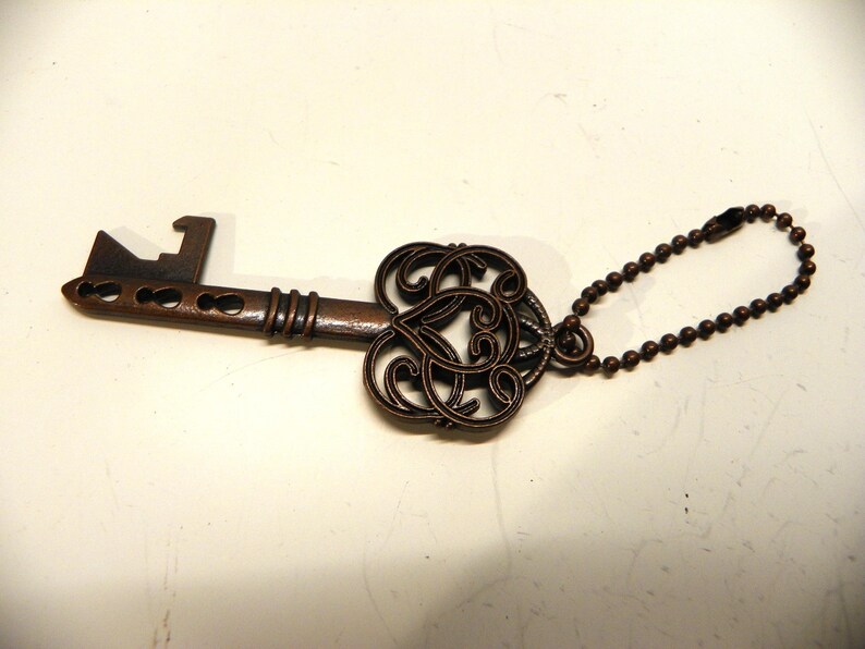 Op de afbeelding: Een koperkleurige metalen flesopener in de vorm van een antieke sleutel. De kop heeft een ingewikkeld opengewerkt krulontwerp en aan de bovenste ring is een klein metalen kogeltjesketting bevestigd om het object op te hangen.
