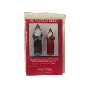 May include: A St. Nicholas Doll craft pattern package featuring two Santa figures, one in a blue coat and one in a red coat. The packaging includes the text St. Nicholas Doll, 2 Santas-2 sizes, 30.48 cm and 35.56 cm high, and Country Appliques.