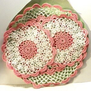 May include: A stack of handmade crochet doilies. Each doily has a central pink circle, surrounded by white and pink crochet patterns. The edges are scalloped with a pink trim. The doilies are set against a green backdrop.