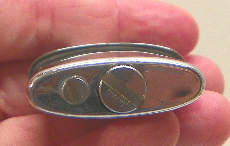 May include: An oval-shaped silver-toned metal lighter base featuring a smooth, reflective surface. The base includes two circular screw-in components, with the larger one clearly engraved with the word JAPAN across its center surface.