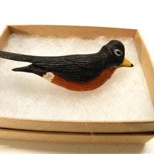以下が含まれることがあります： 黒と白の羽根、赤い胸、黄色いクチバシを持つ手描きの木製ロビン人形。人形は白いティッシュペーパーが入った小さな箱に入っています。