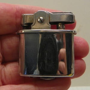 May include: A rectangular silver-toned metal lighter with a polished, reflective finish. The top mechanism features a hinged lever and a central screw, showcasing a classic design with rounded corners and a smooth, metallic surface throughout.