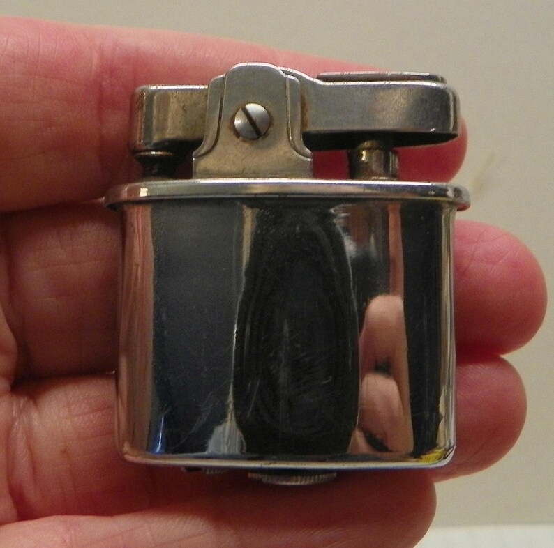 May include: A rectangular silver-toned metal lighter with a polished, reflective finish. The top mechanism features a hinged lever and a central screw, showcasing a classic design with rounded corners and a smooth, metallic surface throughout.