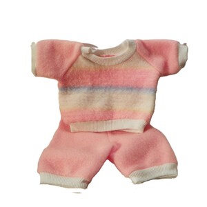 May include: A two-piece outfit consisting of a short-sleeved, fuzzy pink jumper with horizontal pastel stripes in blue, yellow, and white across the chest, paired with matching fuzzy pink shorts featuring white ribbed cuffs at the leg openings.