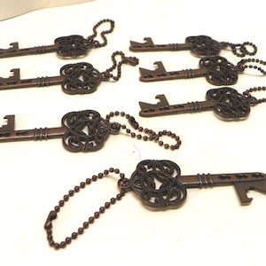 Op de afbeelding: Een verzameling van zes bronskleurige metalen flesopeners in de vorm van een sleutel, elk voorzien van een sierlijke, filigraan-stijl kop en een kogelketting. De sleutels zijn in een groep gerangschikt en tonen hun gedetailleerde koppen en functionele opener-uiteinden.