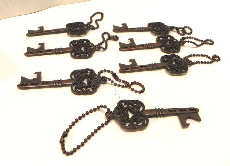 Op de afbeelding: Een verzameling van zes bronskleurige metalen flesopeners in de vorm van een sleutel, elk voorzien van een sierlijke, filigraan-stijl kop en een kogelketting. De sleutels zijn in een groep gerangschikt en tonen hun gedetailleerde koppen en functionele opener-uiteinden.