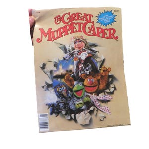 May include: A vintage tan-coloured movie magazine titled The Great Muppet Caper featuring Miss Piggy on a motorcycle, Kermit the Frog, Fozzie Bear, and Gonzo. The cover art includes a blue starburst graphic and a barcode in the bottom left corner.