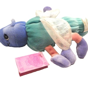 May include: A soft toy insect in shades of blue, teal, pink, and purple. The toy has a smiling face, wings, and a tag that reads "Precious Moments Critter Tails". The toy is designed for children.