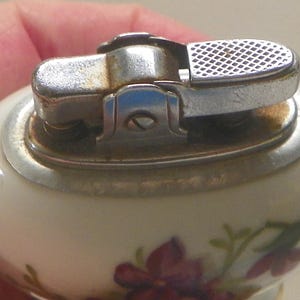 May include: A vintage table lighter with a smooth, off-white ceramic body featuring a floral pattern of purple and green flowers. The top mechanism is made of polished silver-tone metal with a textured ignition lever and a visible screw.