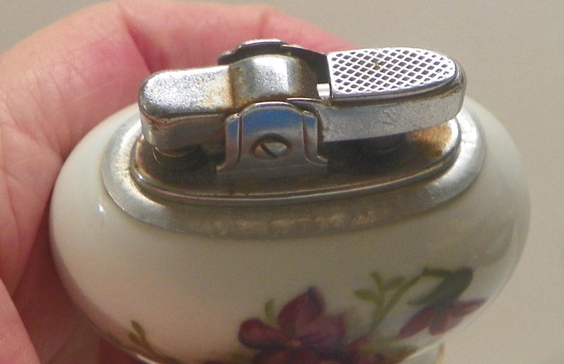 May include: A vintage table lighter with a smooth, off-white ceramic body featuring a floral pattern of purple and green flowers. The top mechanism is made of polished silver-tone metal with a textured ignition lever and a visible screw.