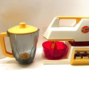 May include: A vintage toy kitchen appliance set featuring a yellow and white stand mixer and a yellow and grey blender. The mixer has a red bowl and a red and white control panel. The blender has a clear container and a yellow lid. The mixer has the text "Lil' Sitter" on the side.