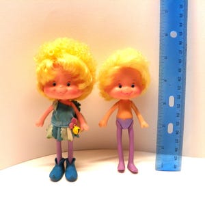 May include: Two vintage yellow-haired dolls with purple and blue clothing. The doll on the left is wearing a blue dress with a flower and bell accessory. The doll on the right is wearing purple trousers and a purple skirt.