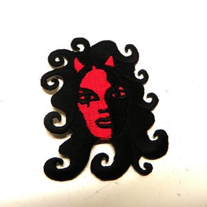 May include: Embroidered patch depicting a stylized face with red skin, black curly hair, and small horns. The patch has a devilish theme, with a detailed design and a bold colour contrast. The patch is likely for clothing or accessories.