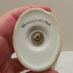 May include: An oval-shaped white ceramic base with a central brass-colored metal screw mechanism. The text "hand painted" is written in black cursive script on the ceramic surface, and the word "JAPAN" is stamped onto the metal screw head.