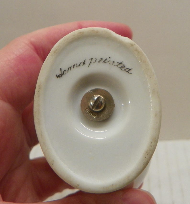 May include: An oval-shaped white ceramic base with a central brass-colored metal screw mechanism. The text "hand painted" is written in black cursive script on the ceramic surface, and the word "JAPAN" is stamped onto the metal screw head.