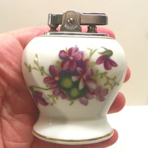 May include: A white ceramic table lighter with a rounded body and a flared base. The surface features a painted floral pattern of purple and pink flowers with green leaves. The top mechanism is made of polished silver-toned metal.