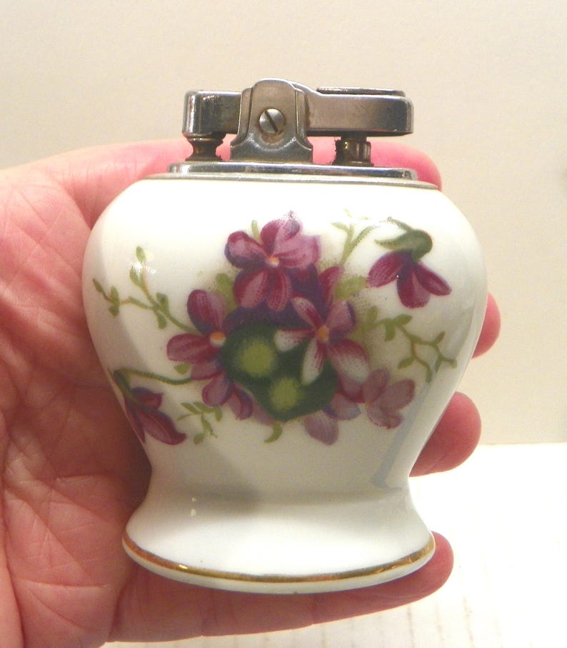 May include: A white ceramic table lighter with a rounded body and a flared base. The surface features a painted floral pattern of purple and pink flowers with green leaves. The top mechanism is made of polished silver-toned metal.