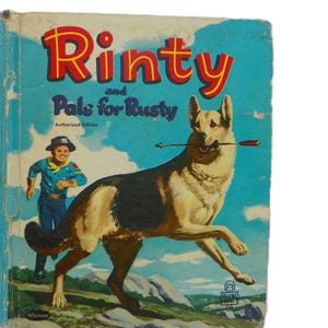 以下が含まれることがあります： 「Rinty and Pals for Rusty」というタイトルのヴィンテージ本の表紙。表紙には、口に矢を持ったジャーマンシェパード、カウボーイの衣装を着た少年、青い空を背景に赤と黄色の文字で書かれたタイトルが描かれています。