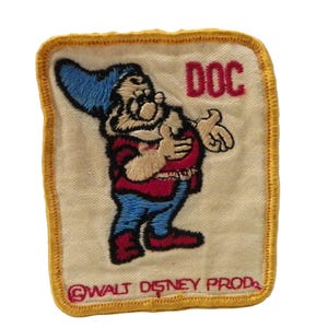 May include: Embroidered patch of Doc from Snow White and the Seven Dwarfs. Doc is shown in his classic attire: blue hat, red vest, and blue trousers. The patch has a yellow border and the text "DOC" and "©WALT DISNEY PROD."