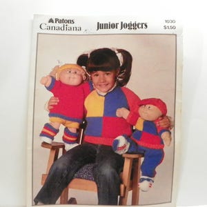 May include: A vintage knitting pattern titled "Junior Joggers" by Patons Canadiana. The pattern shows a child holding two knitted dolls wearing colourful outfits. The dolls are dressed in red, blue, and yellow sweaters and trousers.