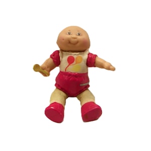 Peut inclure: Une poupée Cabbage Patch Kids vintage, avec une tête chauve, des joues roses et une expression souriante. La poupée porte une chemise blanche avec des graphiques de ballons, une couche rose et des chaussures assorties. Elle tient une cuillère jaune.