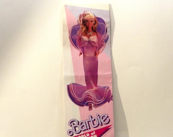 May include: A vintage Barbie World of Fashion promotional card. The card features a Barbie doll in a pink gown with a matching jacket. The Barbie doll has blonde hair and is wearing earrings. The card has a pink and purple color scheme.