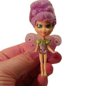 May include: A small fairy doll with vibrant purple curly hair, large green eyes, and a light purple bodice. The doll features translucent pink wings with a swirling pattern and green shoulder accents, standing on small green feet against a white background.