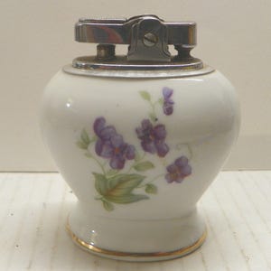 May include: A white ceramic table lighter with a rounded body and a gold-trimmed base. The surface features a painted floral design of purple violets with green leaves. The top mechanism is made of polished silver-toned metal with a textured ignition.