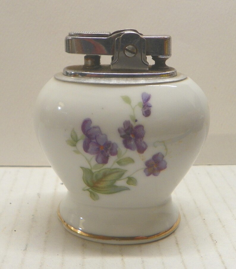 May include: A white ceramic table lighter with a rounded body and a gold-trimmed base. The surface features a painted floral design of purple violets with green leaves. The top mechanism is made of polished silver-toned metal with a textured ignition.