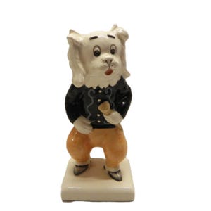 May include: A ceramic figurine of a dog-like character, wearing a black jacket with white trim and orange trousers. The figurine is standing on a rectangular base and holding a small object. The dog has a surprised expression.