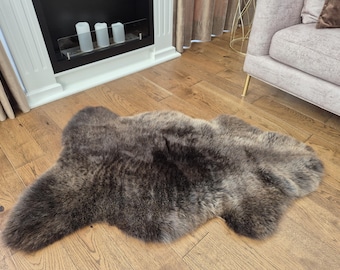 May include: A dark brown sheepskin rug with a natural shape lies on a wooden floor. The rug has a thick, textured appearance with varying shades of brown and gray. The rug is approximately 50 inches long and 30 inches wide.