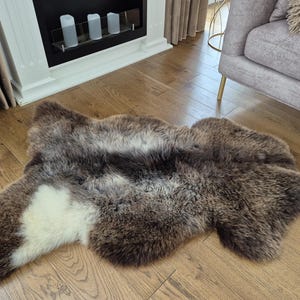Puede incluir: Una alfombra de piel de oveja con tonos marrones, grises y blancos. La alfombra tiene una forma natural e irregular y una superficie gruesa y texturizada. Se coloca sobre un suelo de madera, añadiendo calidez y textura a la habitación.