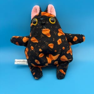 Pode incluir: Um gato de pelúcia preto com manchas laranja. O gato tem olhos amarelos grandes, orelhas rosa e braços estendidos. A etiqueta diz "tumblecosity". Fundo azul.