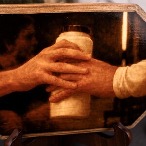 Op de afbeelding: Een sepia-getint kunstwerk dat twee handen toont die een cilindrisch object vasthouden. De handen zijn de focus, waarbij het object een container met een donkere dop lijkt te zijn. Het kunstwerk is omlijst in een houten, achthoekige vorm.