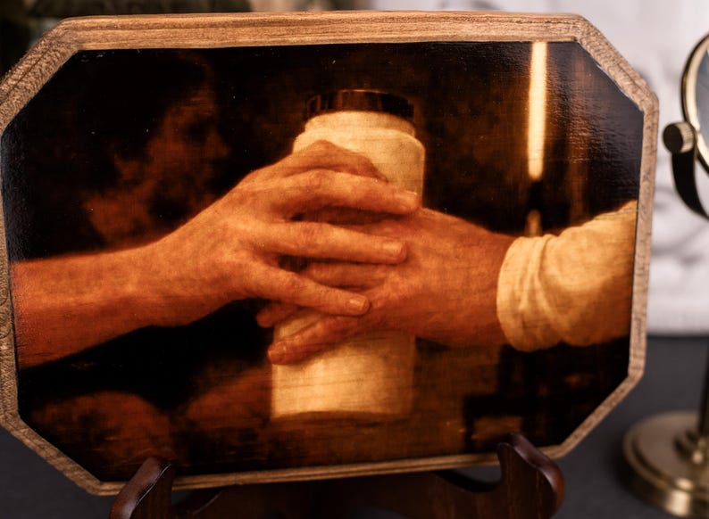 Op de afbeelding: Een sepia-getint kunstwerk dat twee handen toont die een cilindrisch object vasthouden. De handen zijn de focus, waarbij het object een container met een donkere dop lijkt te zijn. Het kunstwerk is omlijst in een houten, achthoekige vorm.