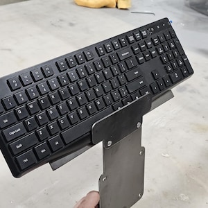 May include: A black computer keyboard with a standard QWERTY layout and a numeric keypad, mounted onto a grey metal bracket. The keyboard features white lettering on each key, including function keys, navigation keys, and a full set of alphanumeric characters.