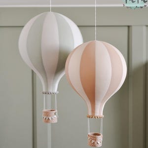 May include: Two decorative hot air balloon ornaments. One is striped in light green and white, the other in peach and white. Each has a woven basket and hangs from a white string. The balloons are suspended against a muted green background.