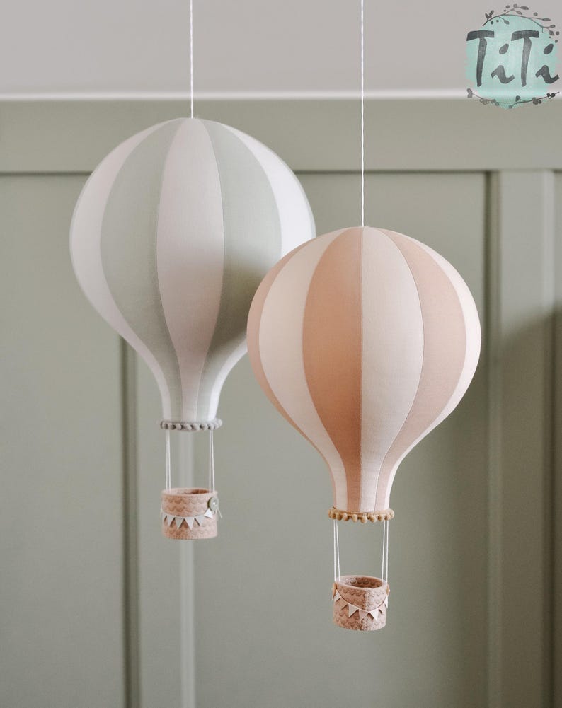 May include: Two decorative hot air balloon ornaments. One is striped in light green and white, the other in peach and white. Each has a woven basket and hangs from a white string. The balloons are suspended against a muted green background.